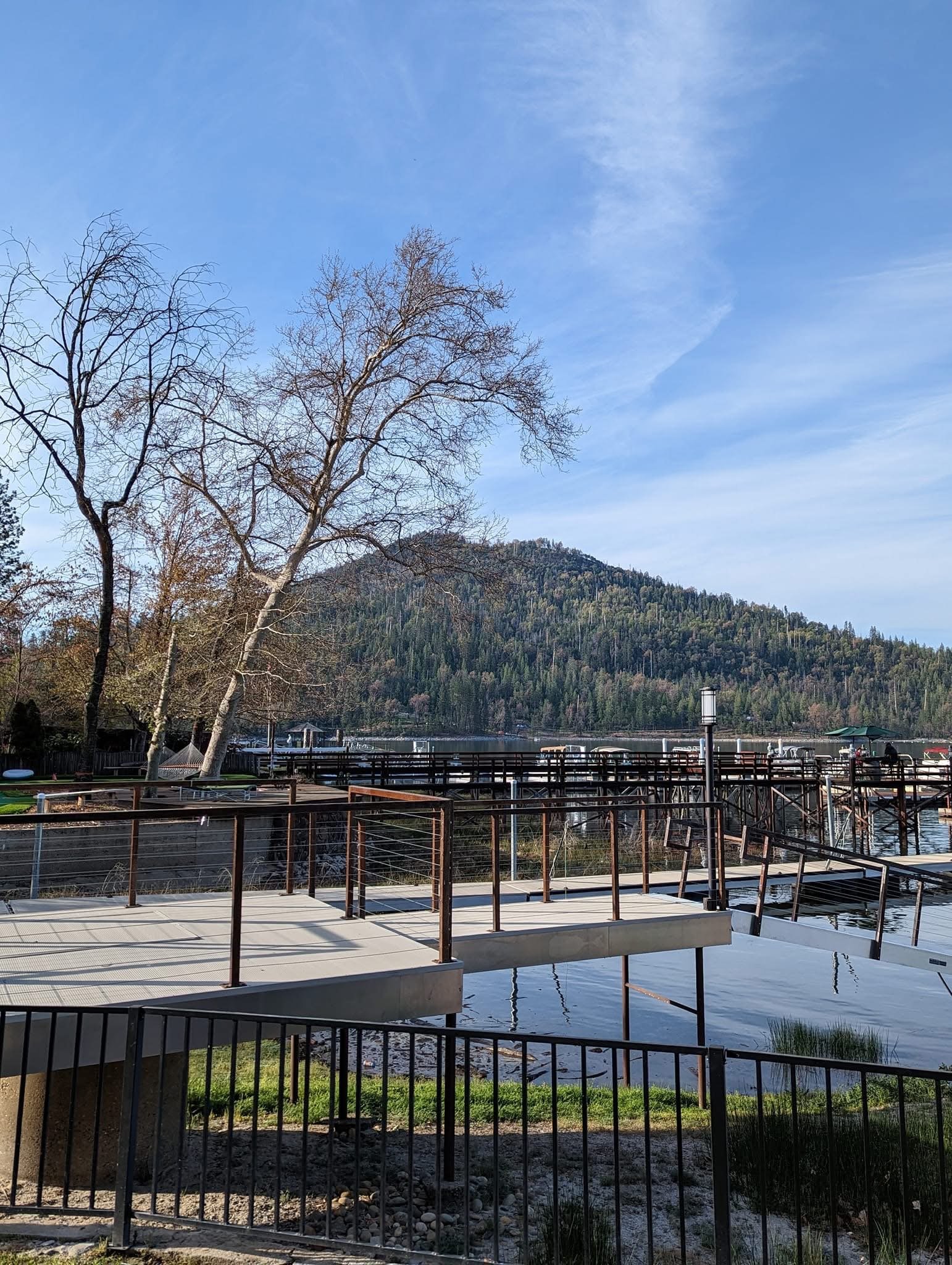 A lakeside scene with a wooden pier and railings, a few leafless trees, and a mountain covered with trees in the background under a partly cloudy sky.