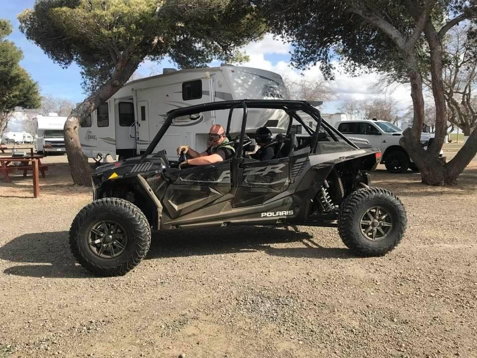 A man and a small dog inside a black Polaris off-road vehicle parked on a dirt lot, with trees and RVs in the background.