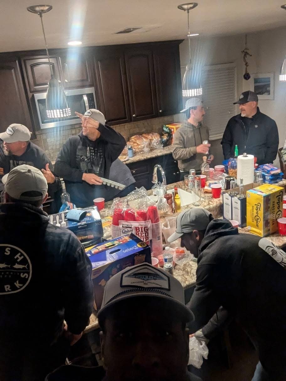 Group of men gathered around a kitchen island, preparing and socializing during a casual get-together with snacks and drinks. The scene appears warm and lively.