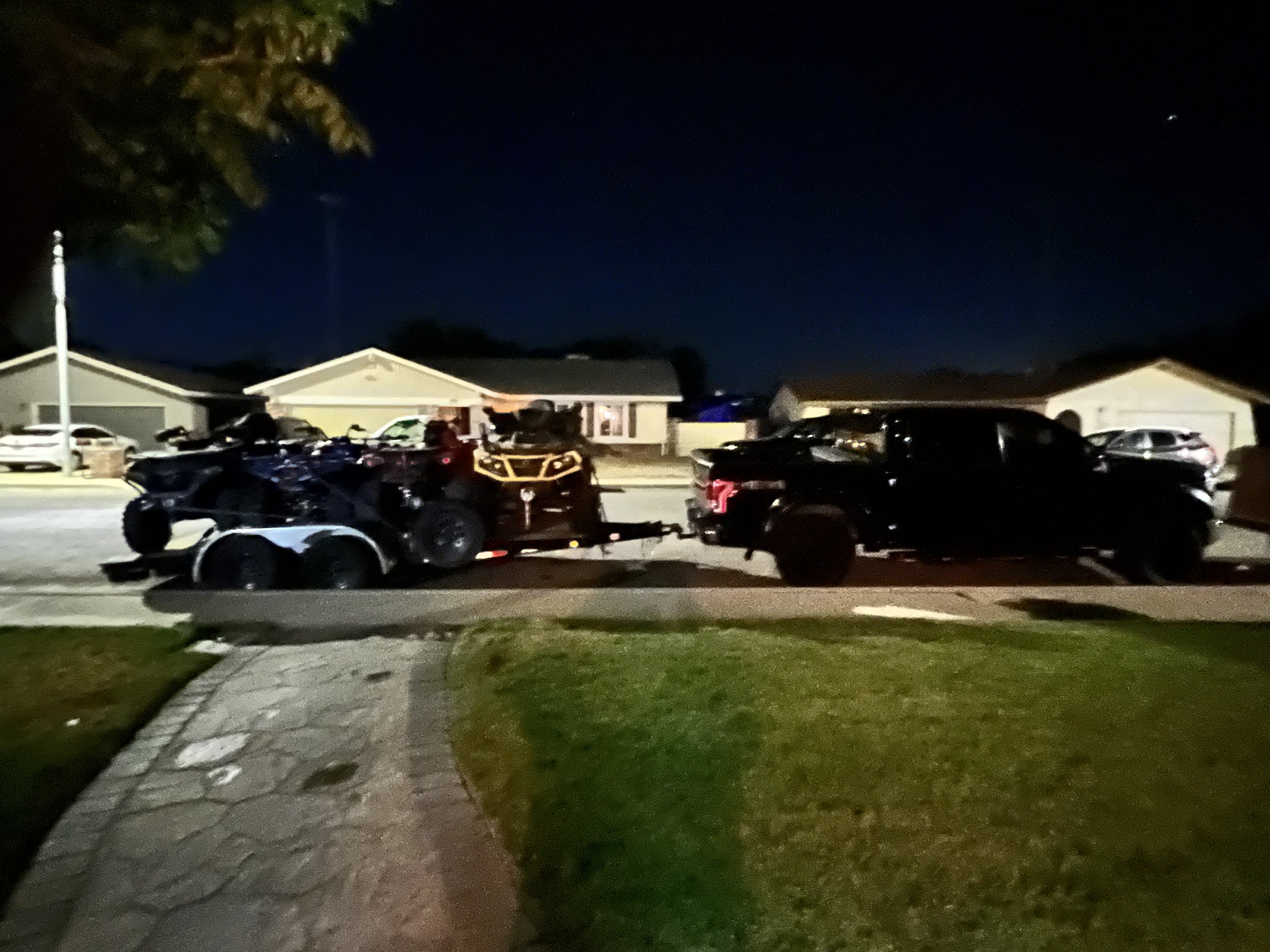 Night scene showing a black pickup truck towing an ATV and a utility terrain vehicle (UTV) on a trailer parked in front of residential houses.