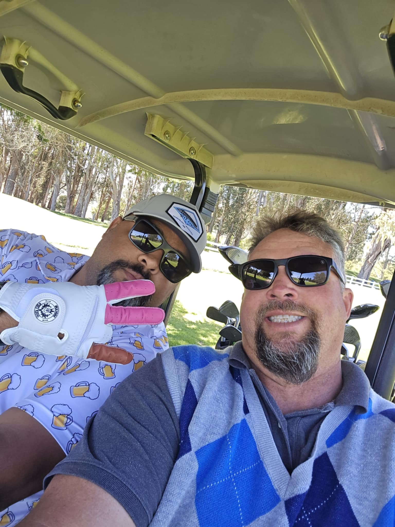 Two men taking a selfie on a golf cart outdoors. One man is wearing sunglasses, a patterned shirt, and a golf glove, making a peace sign. The other man is wearing sunglasses, a gray polo shirt, and an argyle sweater vest. There are trees and a golf c