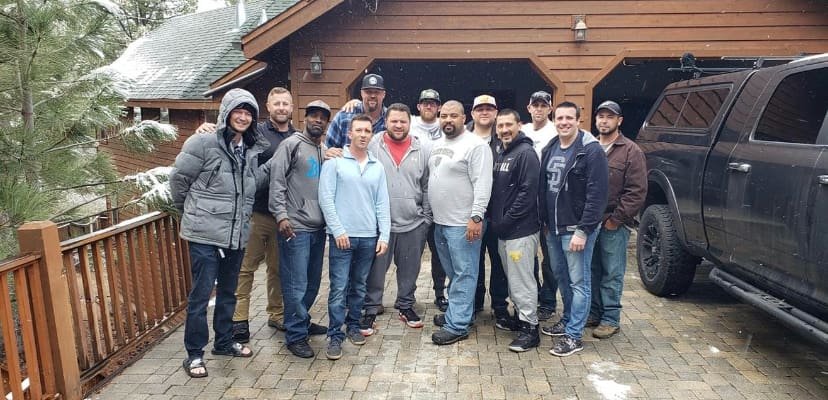 A group of fifteen men standing together outside in front of a wooden building, with a black truck parked nearby on a paved surface, during a rainy day.