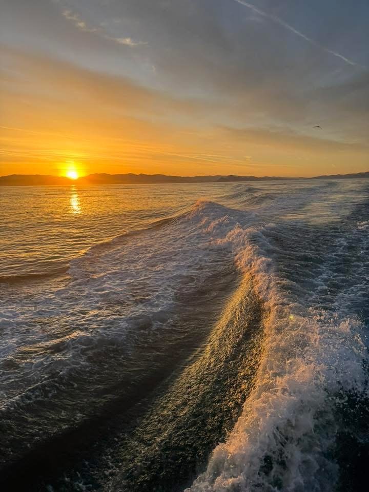 Sunset over the ocean viewed from a moving boat, with waves and water spray in the foreground.