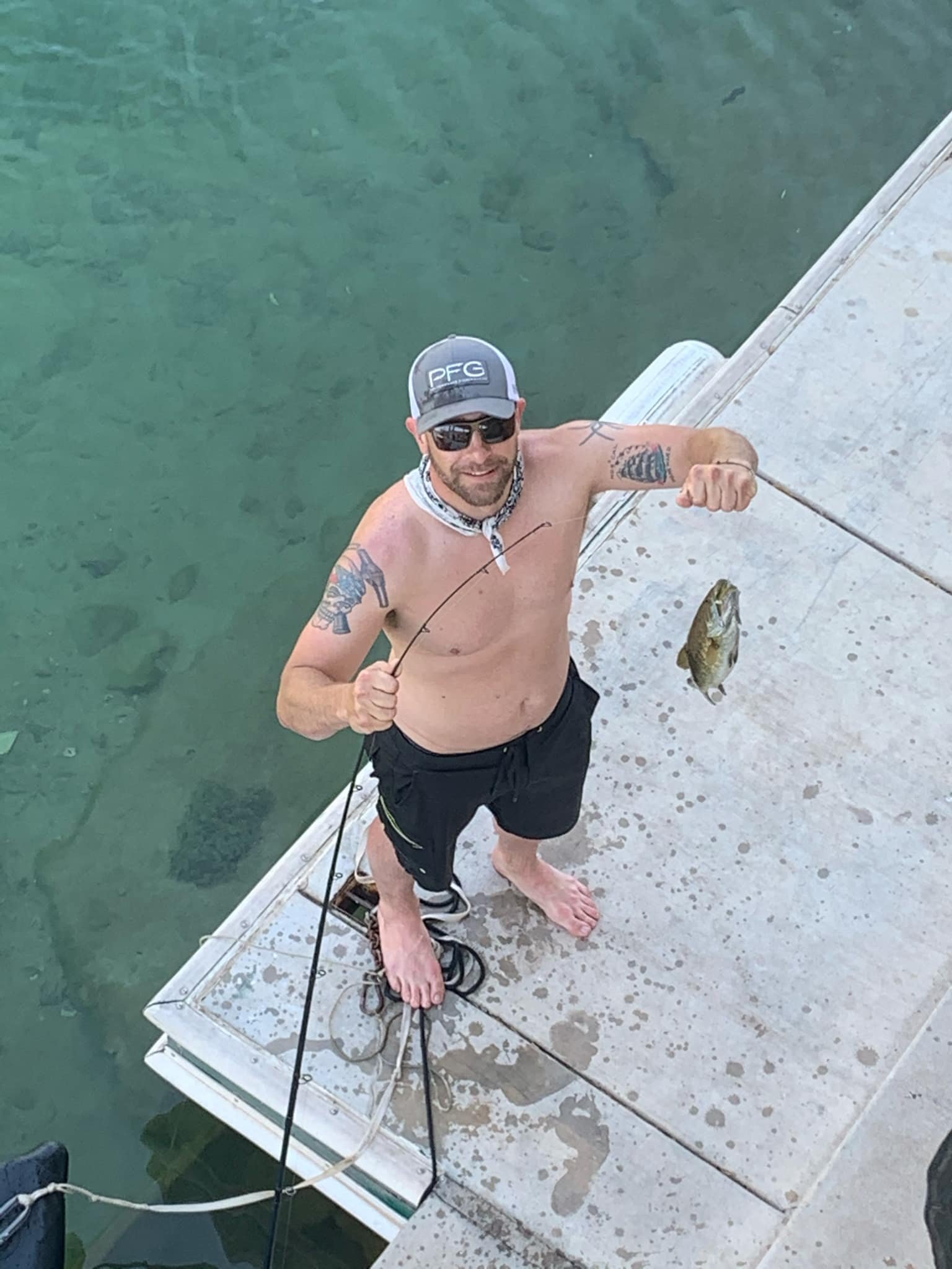 A shirtless man wearing sunglasses, a cap, and black shorts stands on a dock next to green water, proudly holding a fishing rod with a fish he caught.