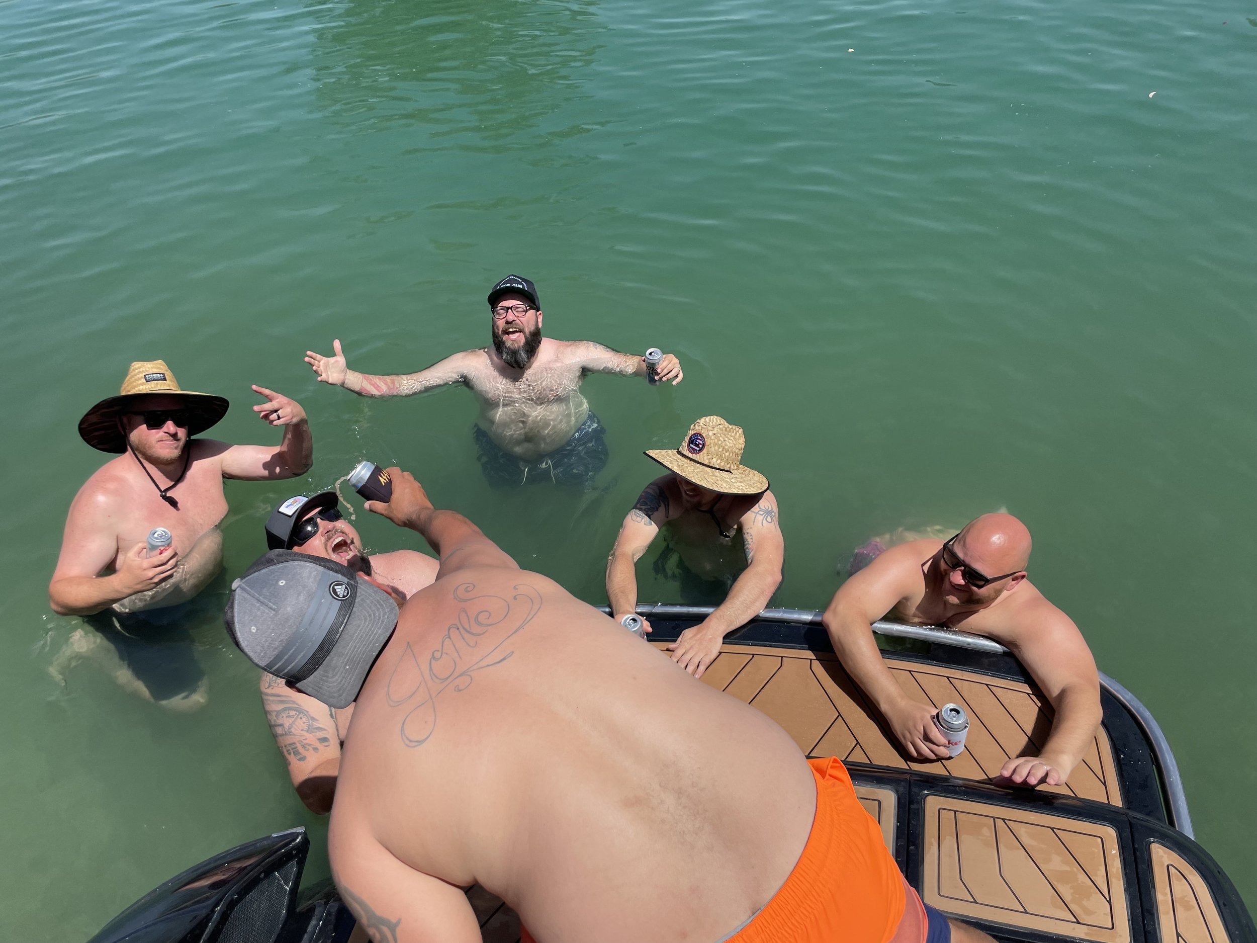 Group of shirtless men, some wearing hats and sunglasses, relaxing in green water, with a wooden deck visible at the bottom, holding cans and smiling.