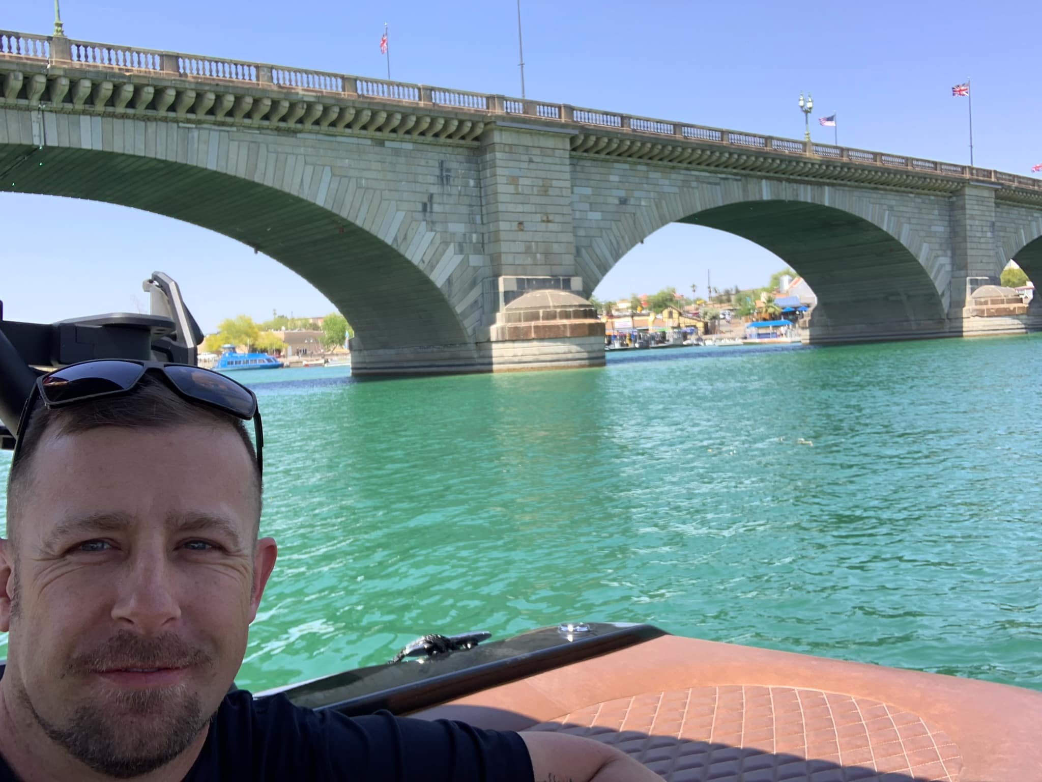 A man with sunglasses on his head taking a selfie on a boat with a bridge and water in the background.