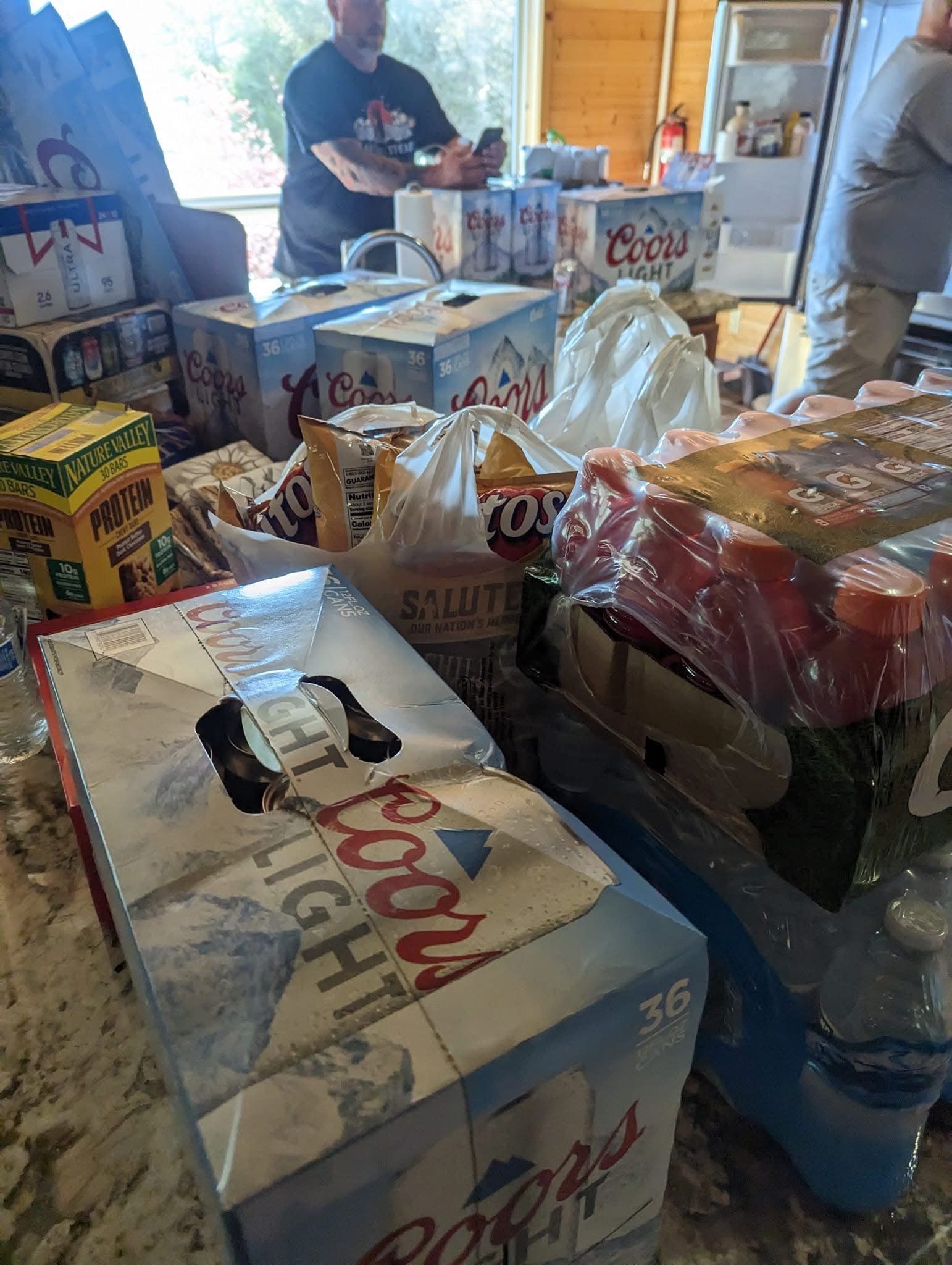 A table filled with grocery items including beer cases, snack bags, bottled water, and other packaged foods, with two people in the background near a window and an open refrigerator.