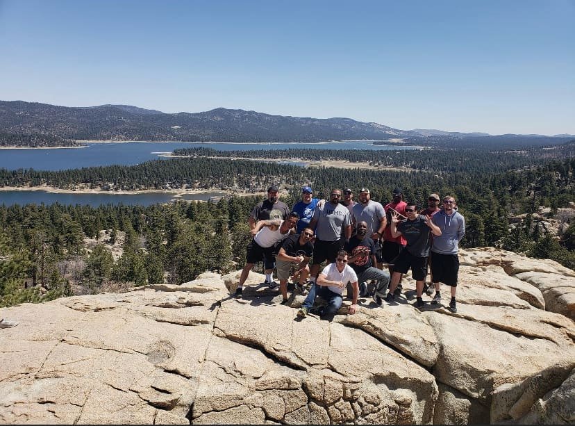 Group of ten friends posing on a large rock formation overlooking a scenic lake and forested mountains.