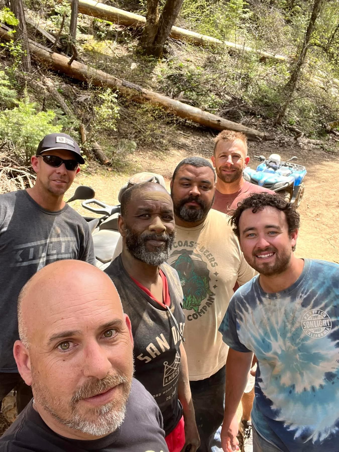 Six men standing together in a wooded outdoor area with two ATVs visible in the background.