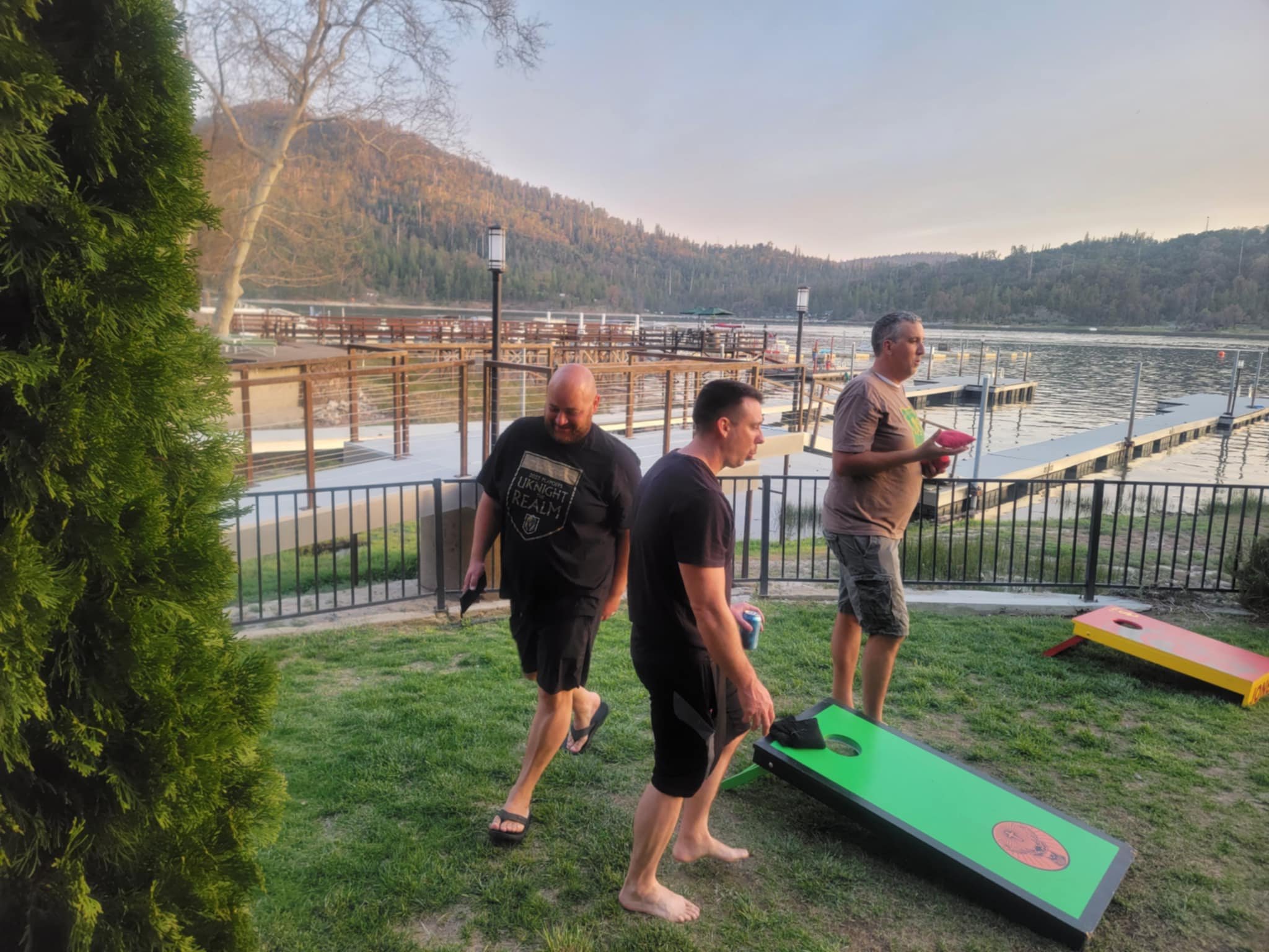 Three men playing cornhole outdoors by a lake with a dock and mountains in the background, during sunset.