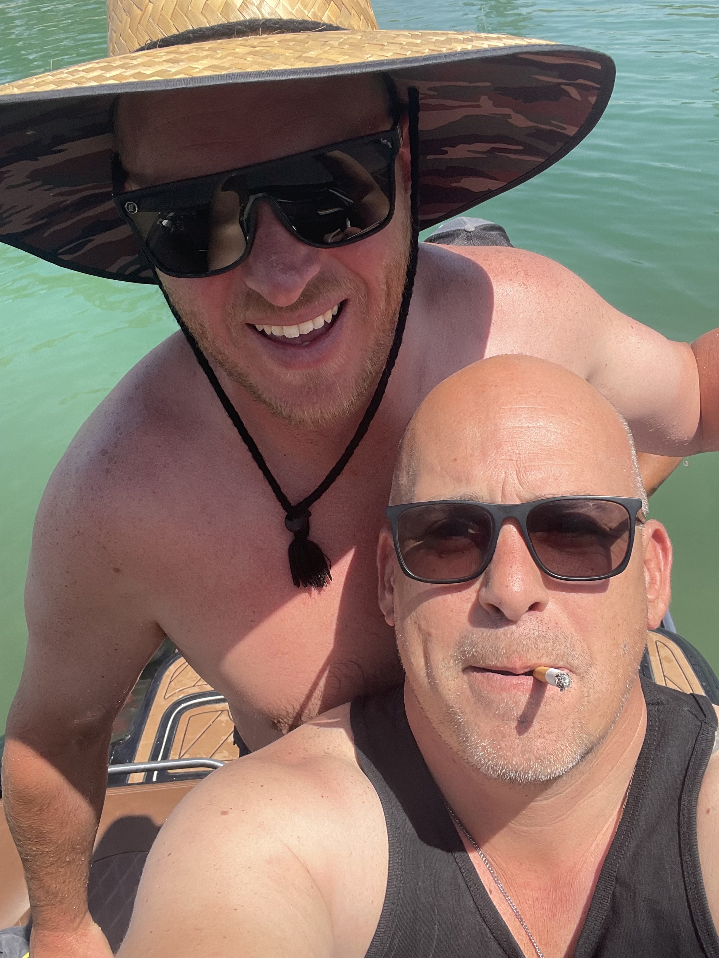 Two men with sunglasses on a boat, one with a cigarette and a hat, smiling, in a body of water on a sunny day.