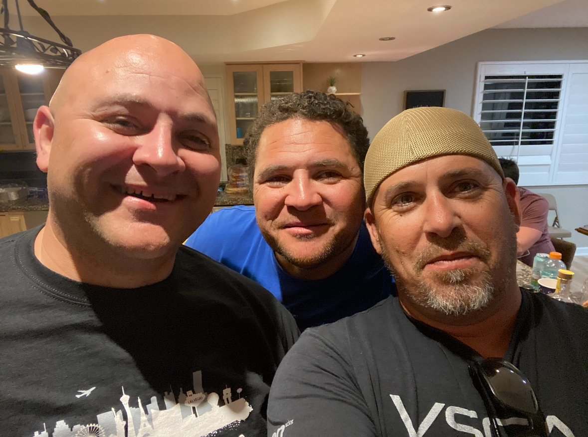 Three men taking a close-up selfie in a home kitchen with cabinets, a window with shutters, and a person sitting at a table in the background.