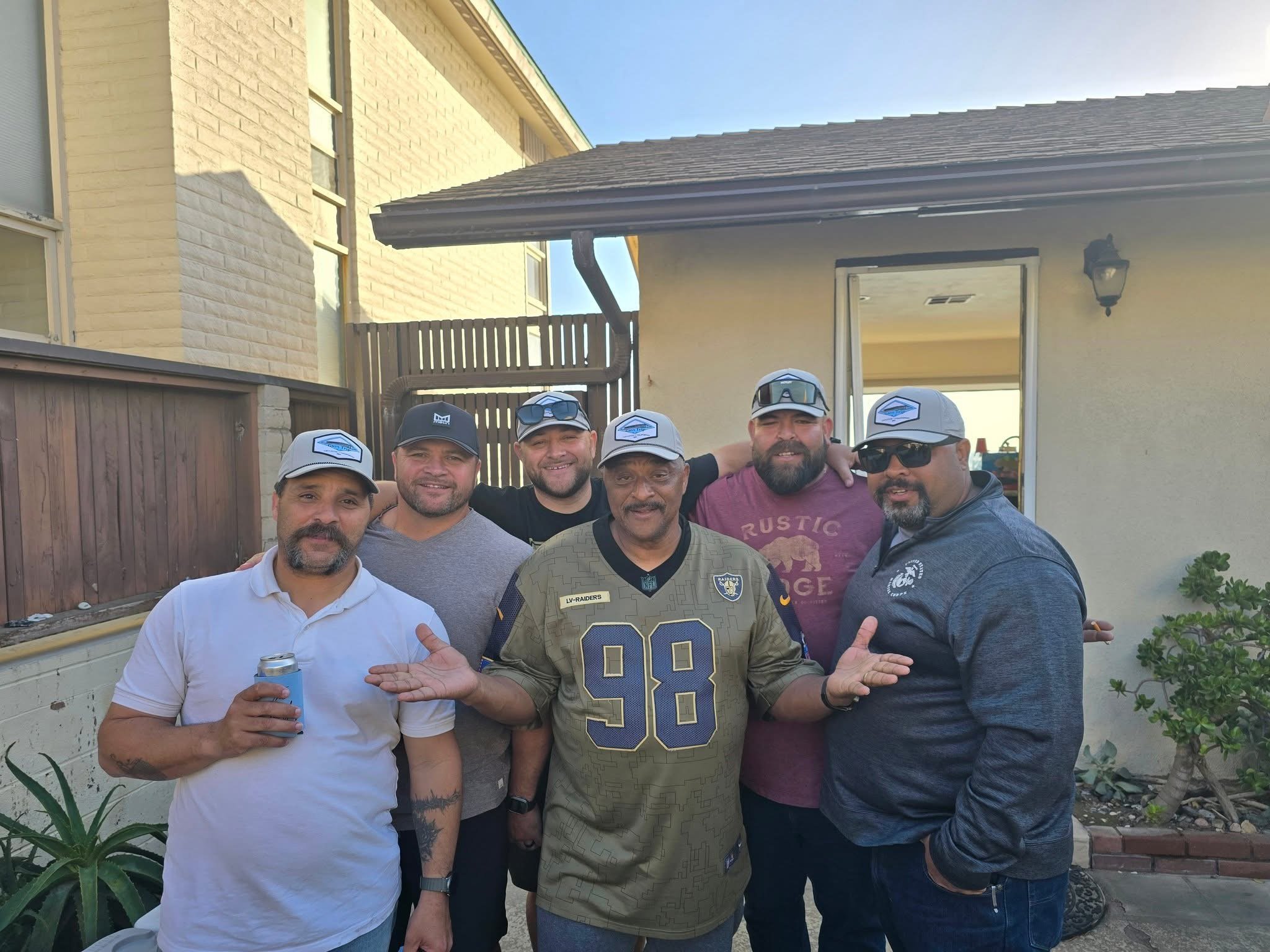 Six men, some wearing gray hats with a logo, standing outdoors in front of a house, smiling, with some holding drinks, and one man wearing an Oakland Raiders football jersey.