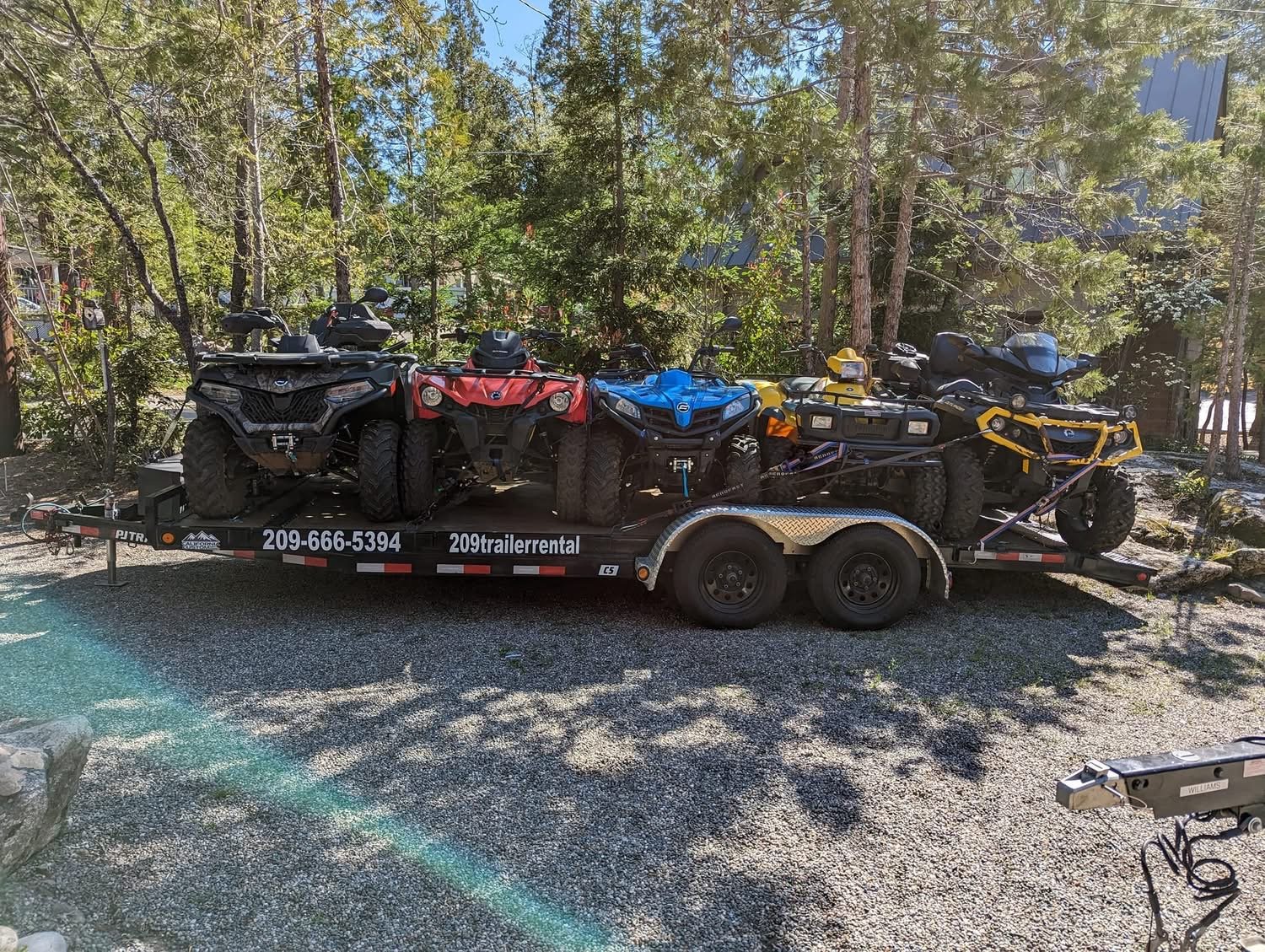 A trailer loaded with six all-terrain vehicles (ATVs) parked on gravel with trees and a building in the background.