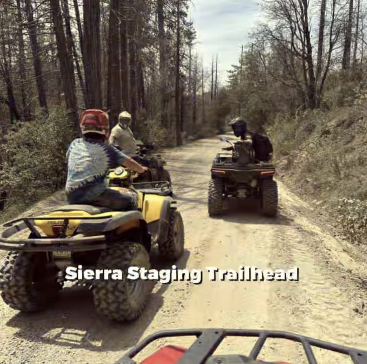 Three people riding all-terrain vehicles on a dirt trail in a forested area with trees and bushes, with the text 'Sierra Staging Trailhead' overlaid.