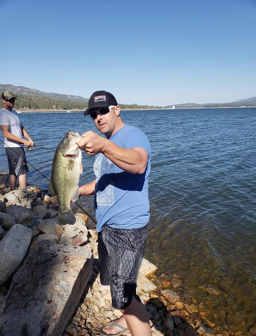 Man wearing sunglasses, a black cap, and a blue T-shirt holding a large fish on a rocky lakeshore with another person fishing in the background.