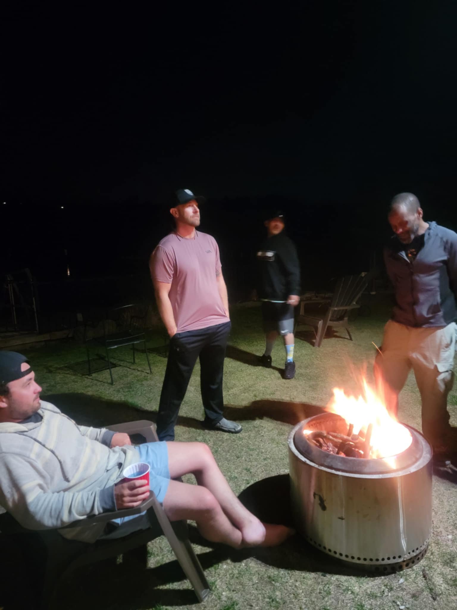 Four men gathered outdoors at night, some standing and one sitting in a lawn chair, around a lit fire pit on a grassy area. One man is sitting, holding a red cup, and the others stand nearby.