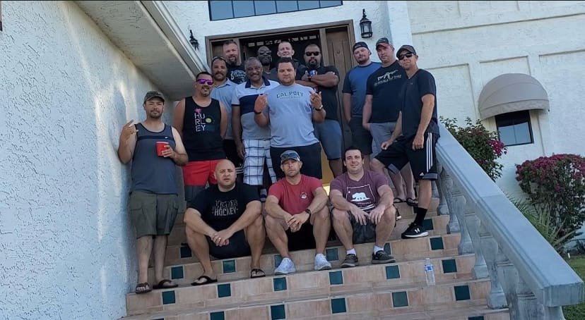 A group of 13 men gathered on the steps of a house for a photo. They are casually dressed, some holding drinks, with one man in a California Police shirt. The house has a white exterior, wooden door, and flower bushes nearby.