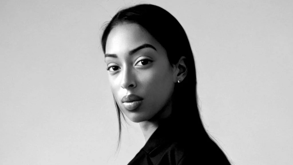 A black and white portrait of a young woman with long, straight hair, wearing earrings, and looking directly at the camera against a plain background.