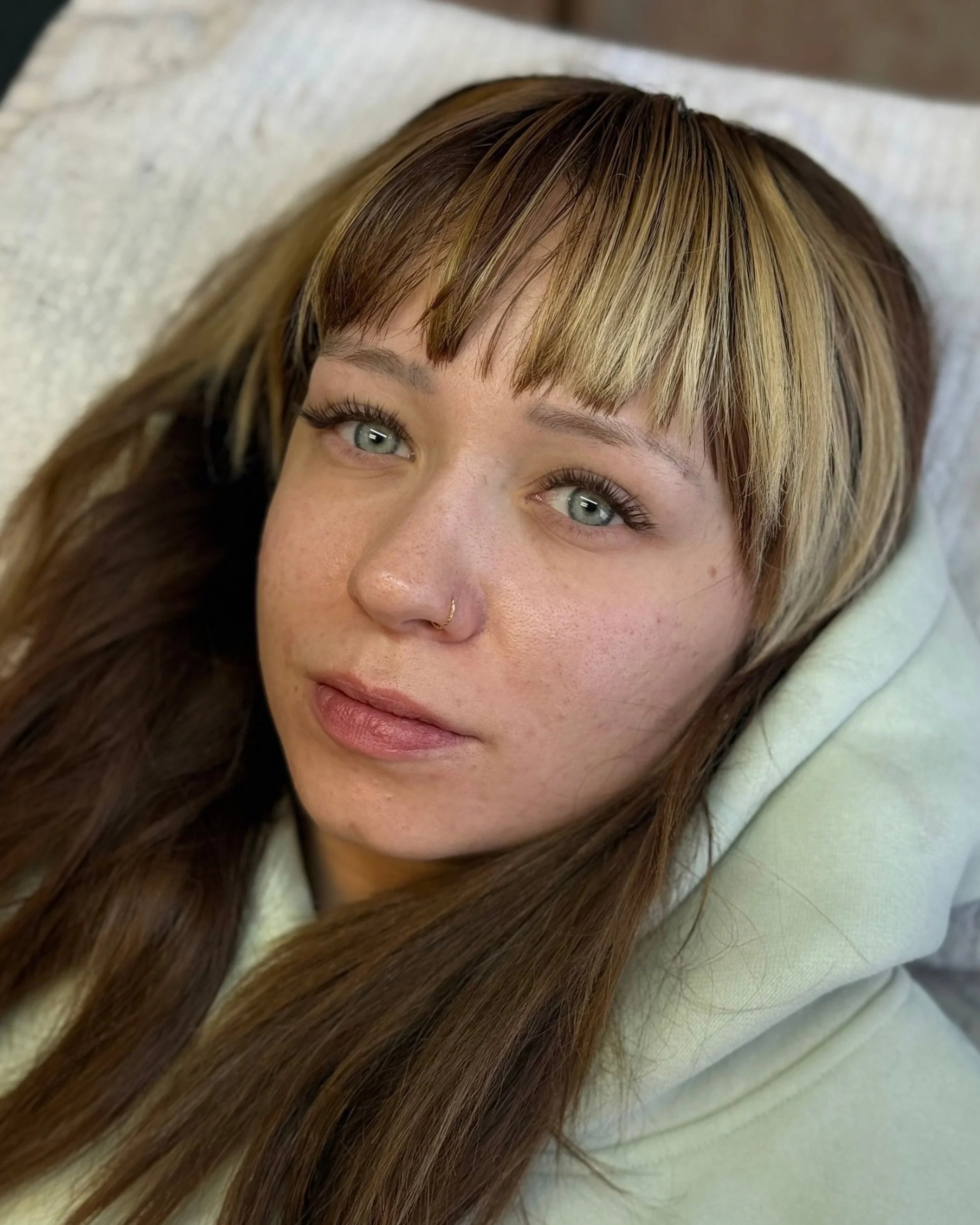 Close-up of a woman with light skin and multiple shades of brown and blonde hair, lying on a cream-colored surface, looking directly at the camera.