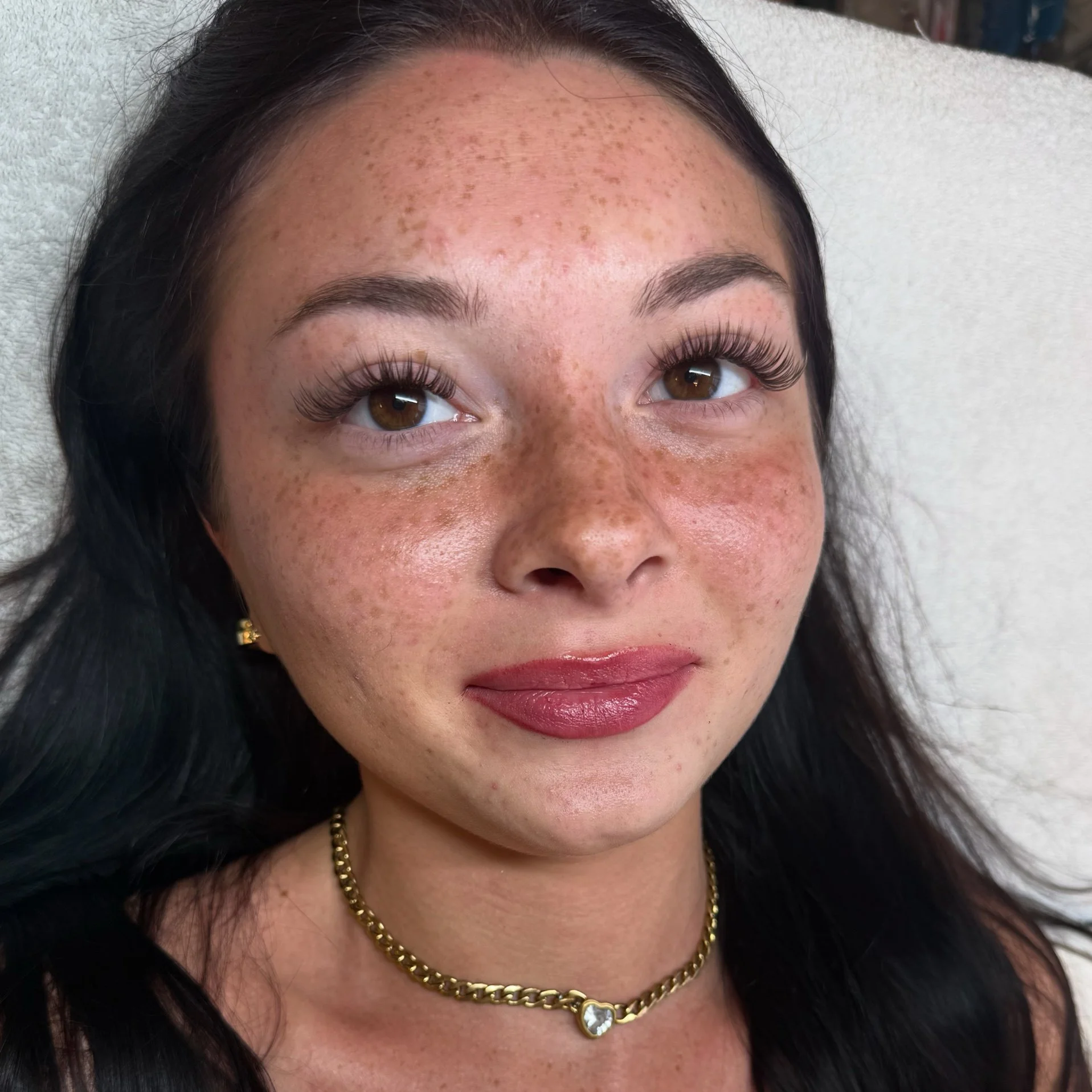 Close-up of a woman's face showing her forehead with freckles and her dark hair, and part of her lips with natural makeup, she is wearing a gold necklace with a heart-shaped pendant.
