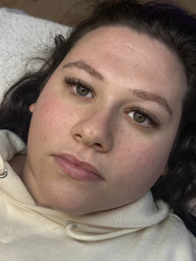 Close-up of a young woman's face with light skin, hazel eyes, dark eyebrows, and dark hair, lying down and wearing a cream-colored hoodie.