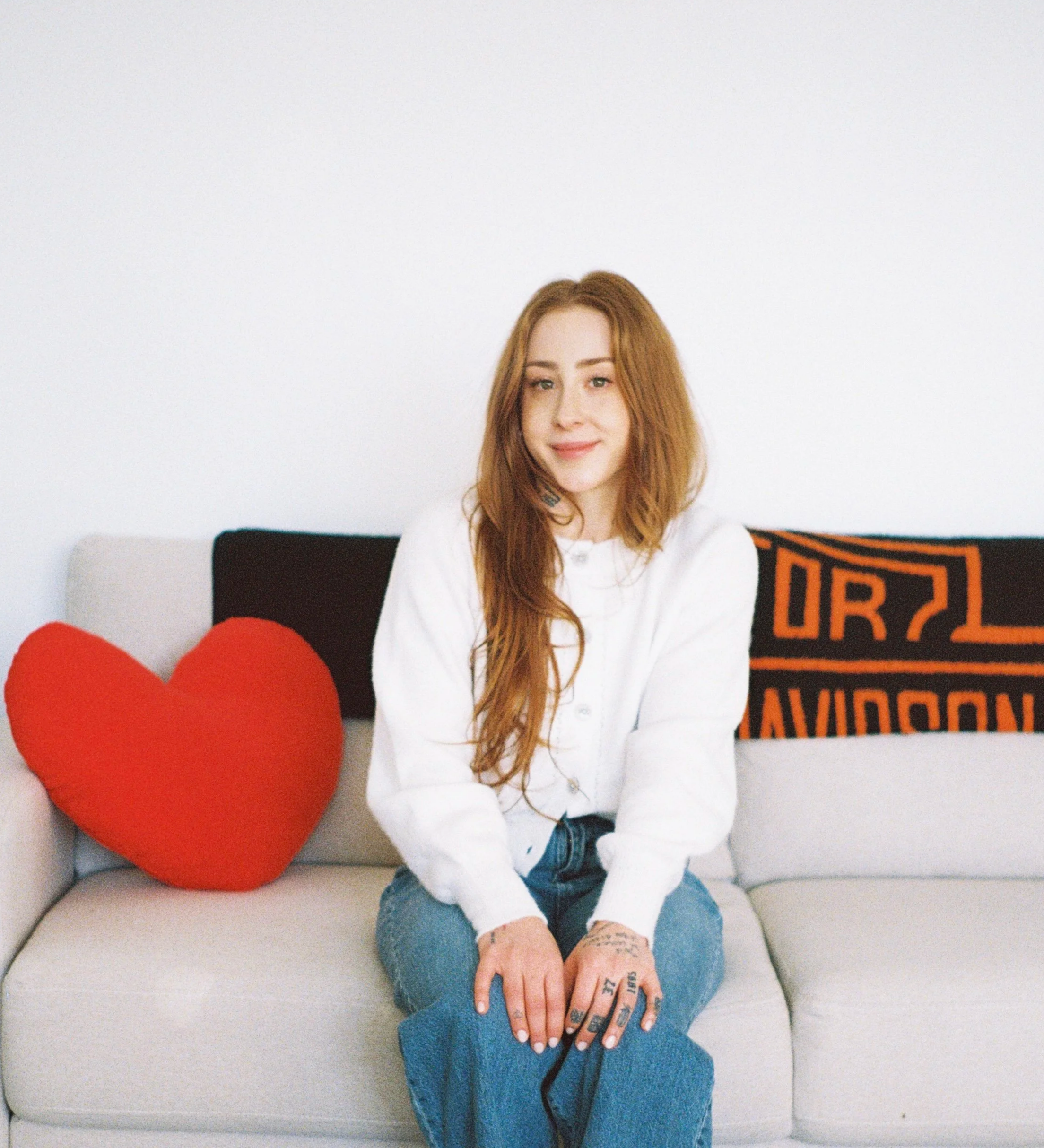 A woman with long red hair sitting on a beige sofa with a large red plush heart and a framed sign in the background that reads "URBAN." She is wearing a white shirt, ripped blue jeans, and has tattoos on her hands and fingers.