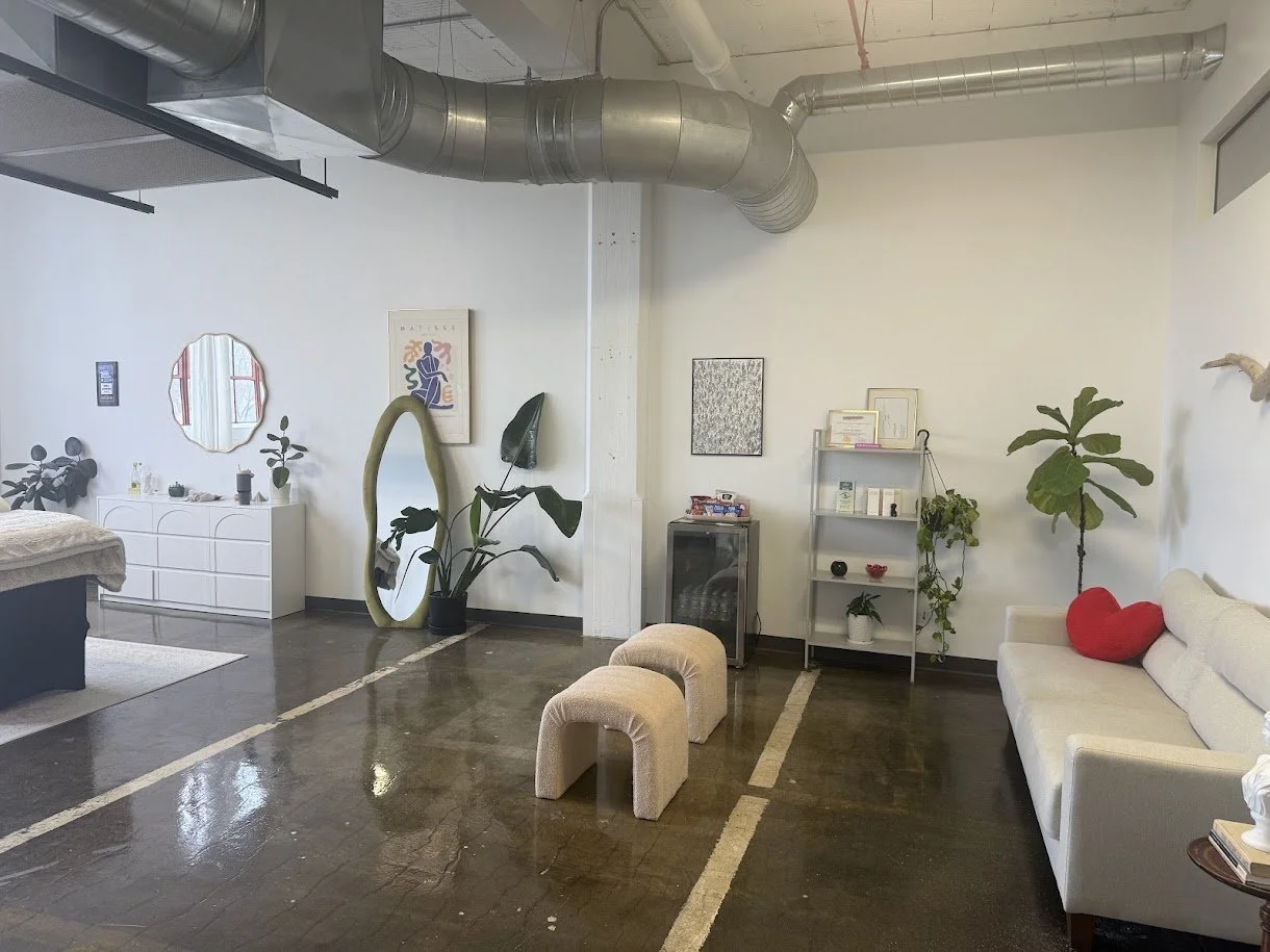 A modern, minimalist living room with white walls, industrial ductwork ceiling, dark wood floor, and various furniture including a couch, small stools, a tall mirror, potted plants, shelves, and wall decorations.