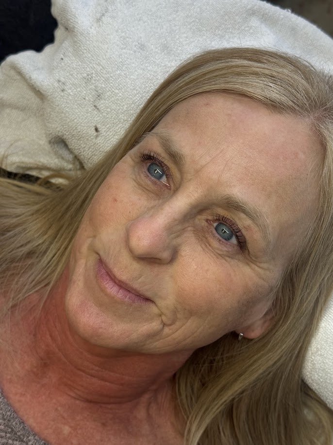A close-up photo of a middle-aged woman with blonde hair and blue eyes, resting on a white towel or blanket.