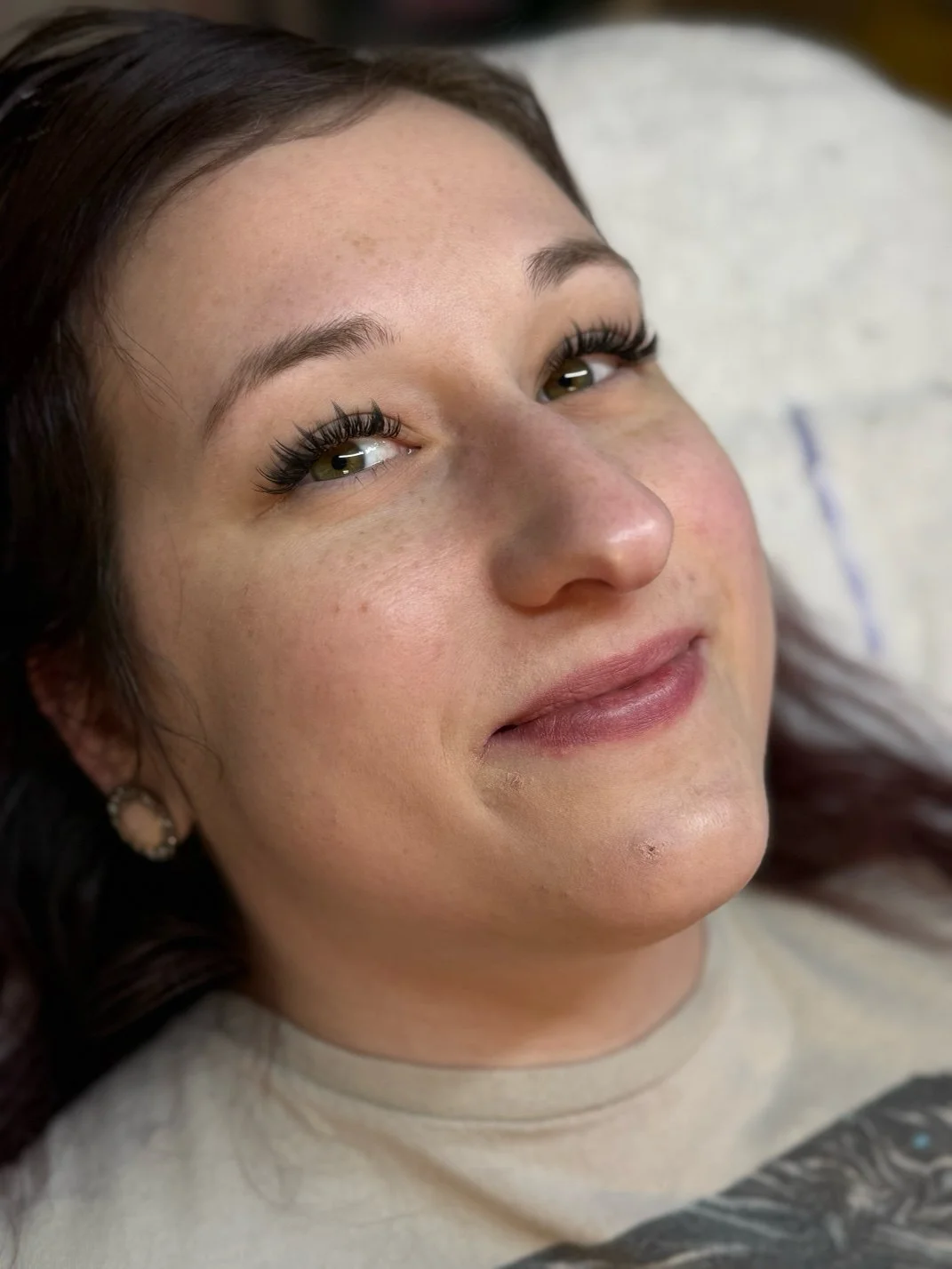 A woman with dark brown hair, lying down and smiling softly, with natural makeup and long eyelashes.