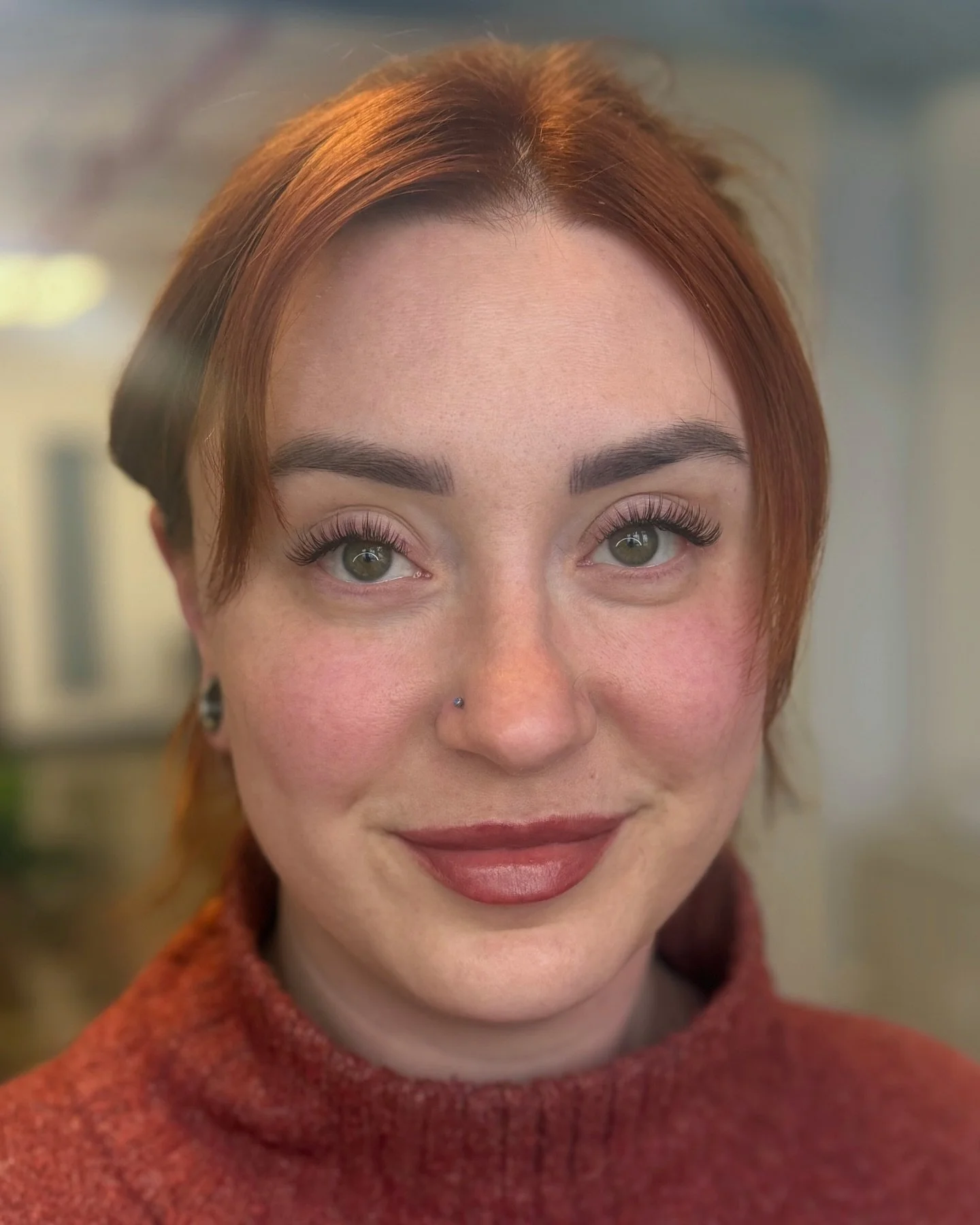 Close-up portrait of a smiling woman with red hair, green eyes, and a nose piercing, wearing a red sweater, in an indoor setting.