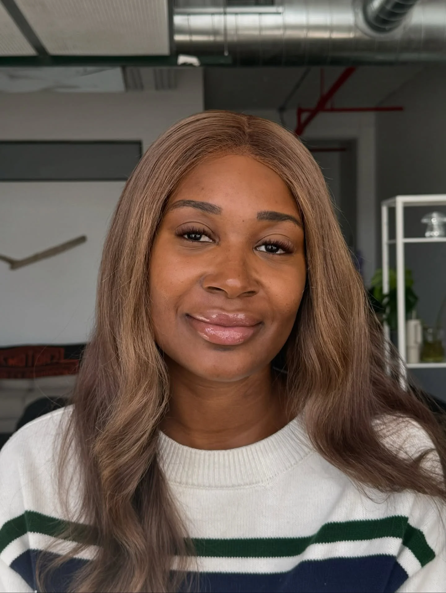 Close-up of a woman with long, wavy, light brown hair, wearing a white sweater with green, navy blue, and gray stripes, smiling softly, in an indoor setting with industrial ceiling pipes and a white shelving unit in the background.