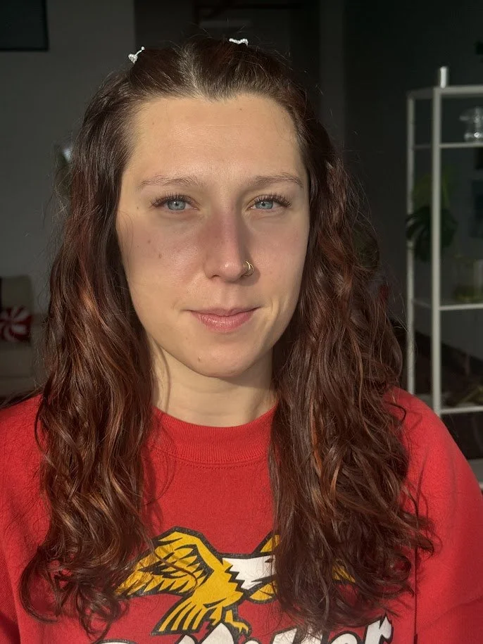 A young woman with long, curly brown hair, light skin, and blue eyes, wearing a red shirt with a yellow and black eagle logo, standing indoors near a shelving unit and a plant.