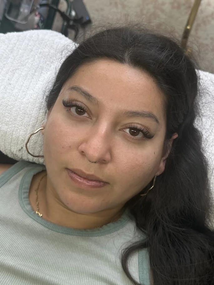 Close-up of a young woman with dark hair, lying on a white pillow, wearing eyelashes, gold hoop earrings, a gold necklace, and a septum piercing, with a blurred background.