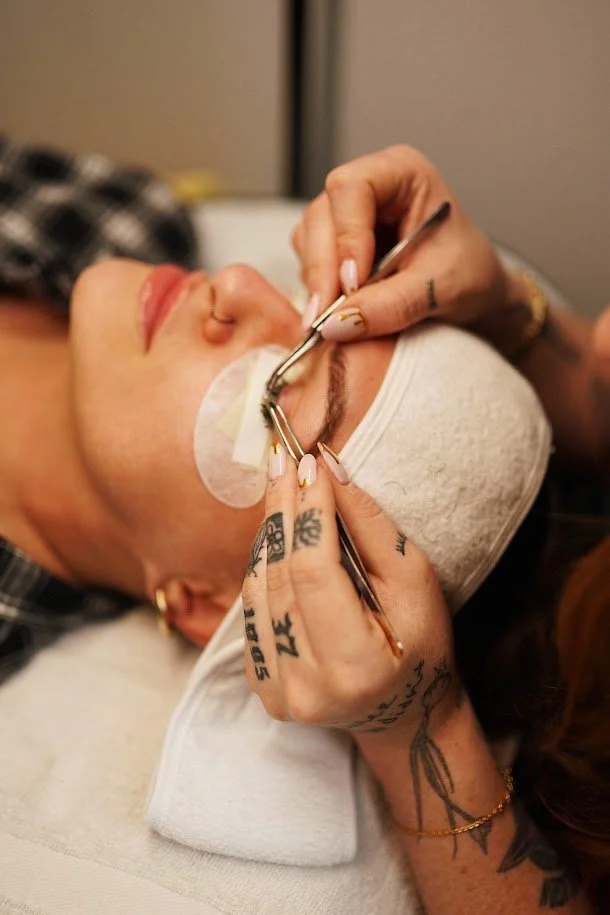 A woman with tattoos receiving a cosmetic eyebrow procedure with tweezers and a small tool, lying down with a towel over her head and protective eye patch.