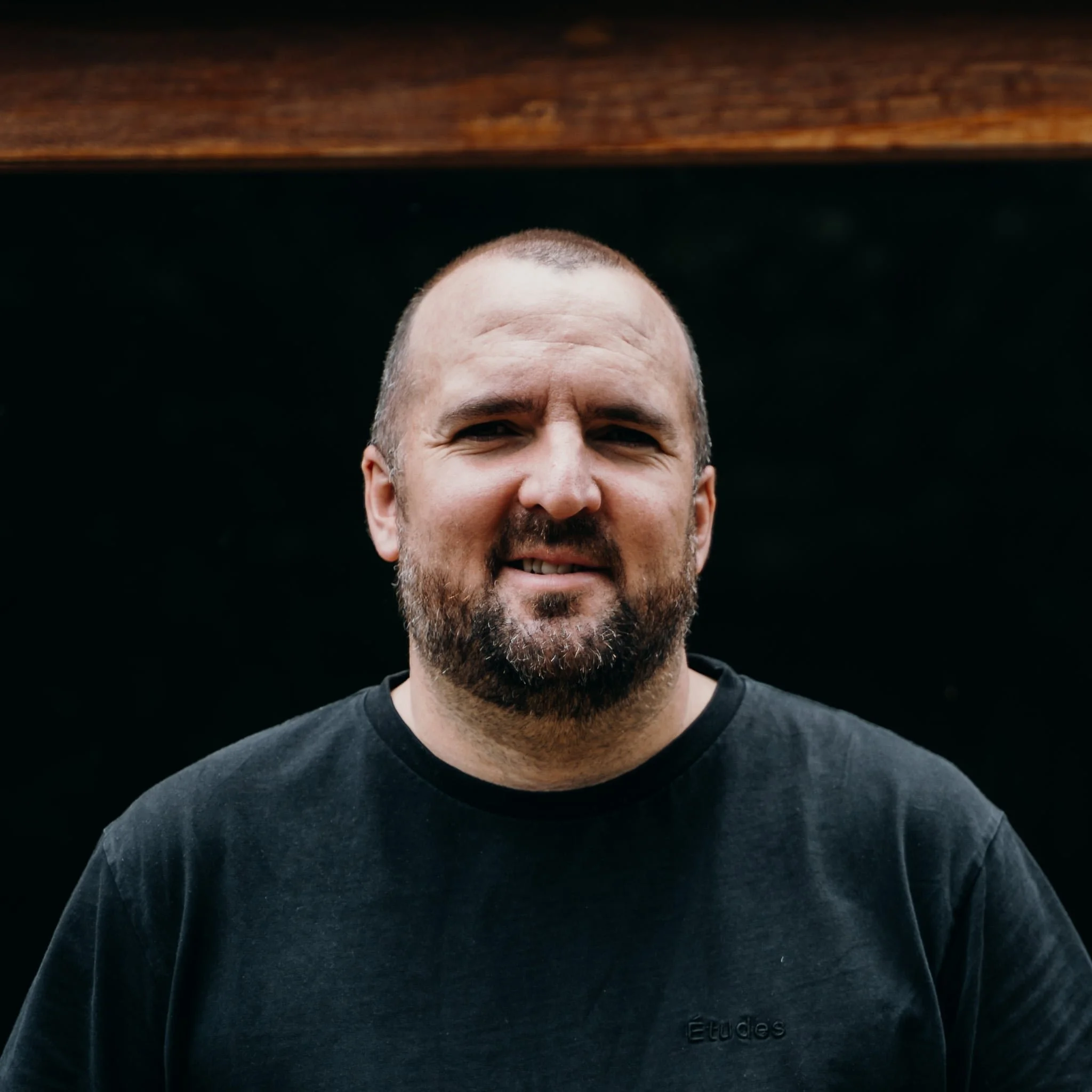 Portrait of a man with a beard, short hair, wearing a black T-shirt, standing outdoors against a dark background.