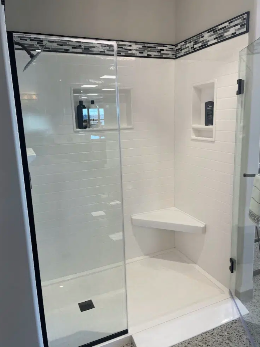 A modern, white-tiled shower with a clear glass door, a built-in niche with shampoo bottles, a small corner bench, and a decorative mosaic tile border at the top.