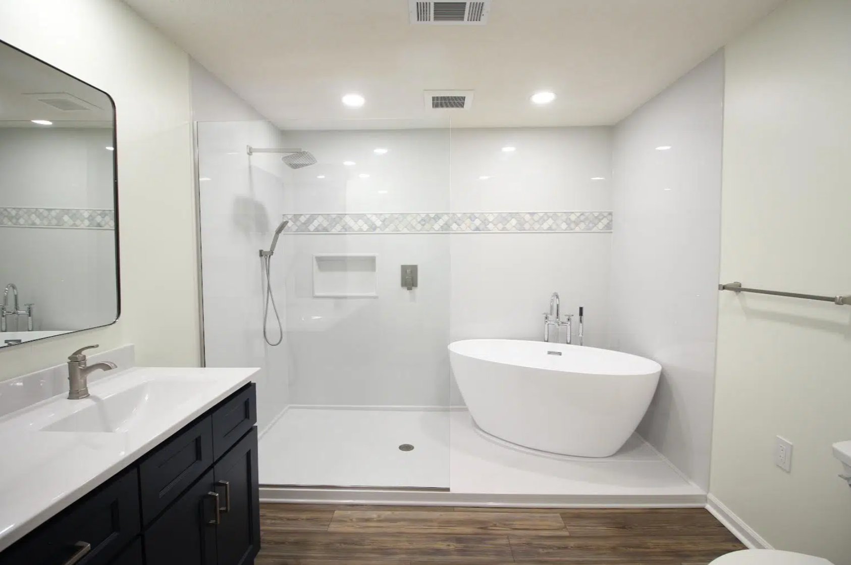 Modern bathroom featuring a vanity with a white countertop and dark cabinets, a large mirror, a walk-in shower with a rainfall showerhead, and a freestanding bathtub, with wood flooring.