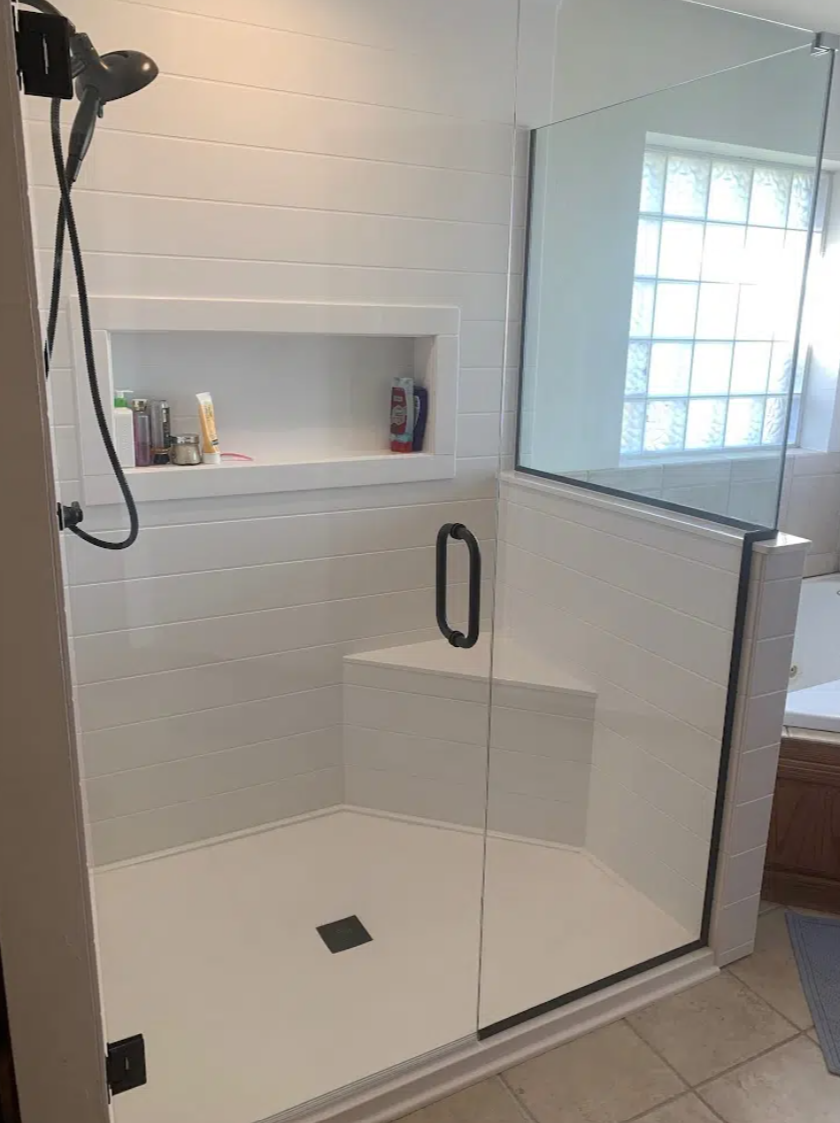 A modern walk-in shower with clear glass walls, a black handle, white tiled walls, a built-in shelf with toiletries, a black showerhead, and a window letting in natural light.