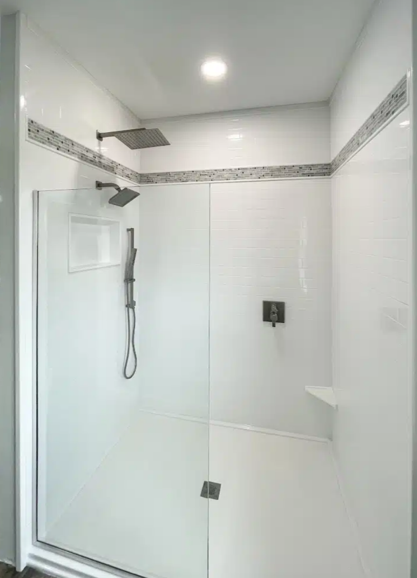 Modern walk-in shower with a large rainfall showerhead, a handheld shower, a small built-in shelf, and white tile walls with decorative border.