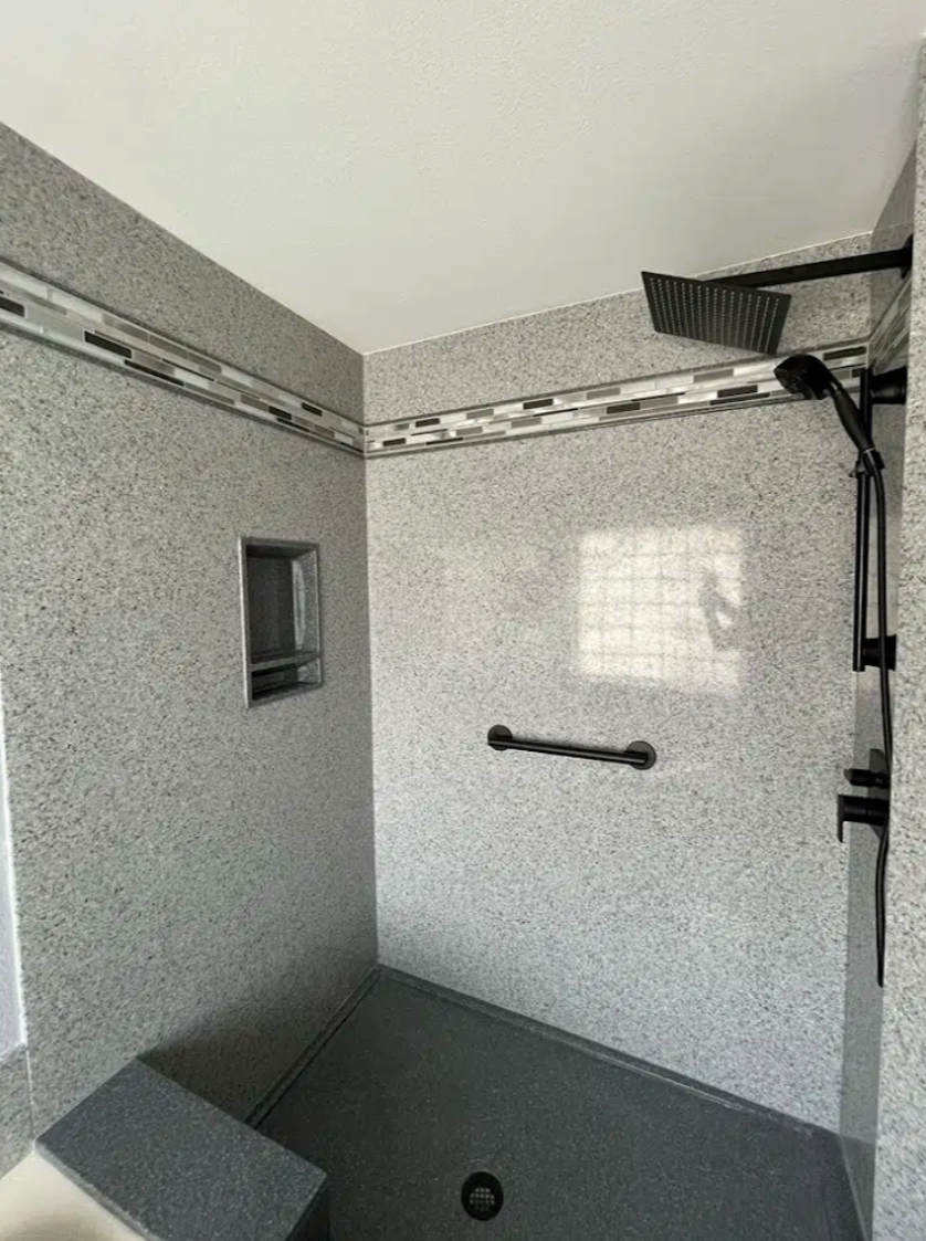 Accessible walk-in shower with gray speckled walls, a small built-in shelf, a grab bar, a rainfall showerhead, and a textured, non-slip floor.