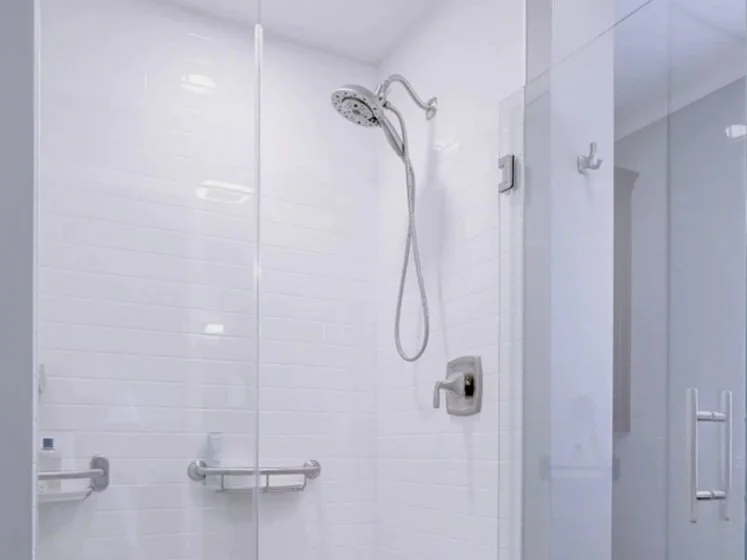 Shower area with a rainfall showerhead, a handheld shower, white tiled walls, safety bars, and a glass door.