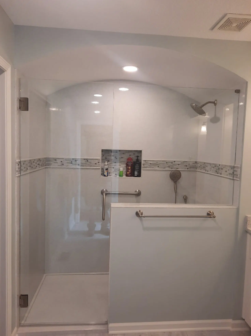 A walk-in shower with glass doors, a built-in niche for toiletries, a handheld showerhead, an overhead shower, and safety grab bars in a bathroom.