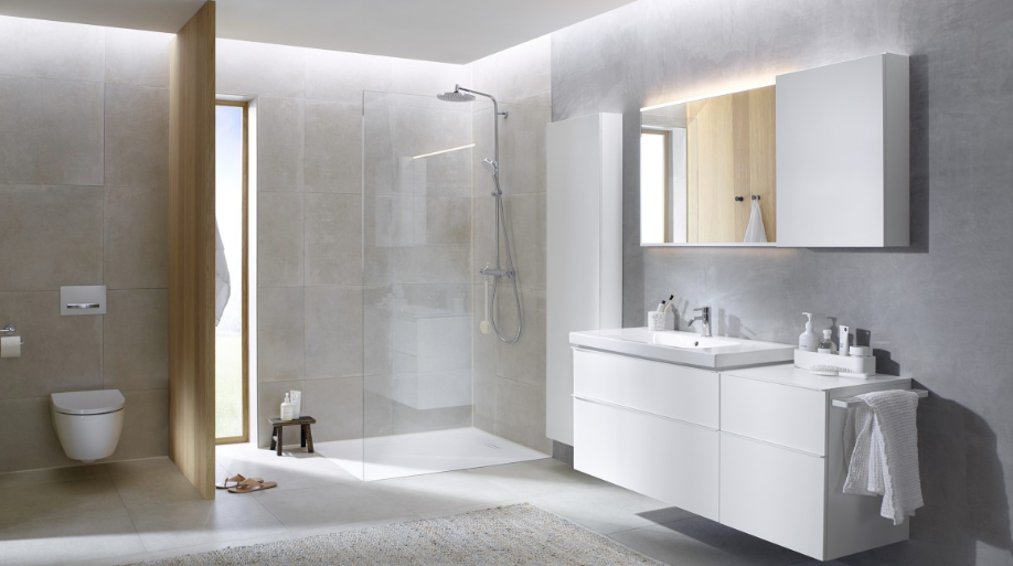 Modern bathroom with a walk-in glass shower, white vanity with toiletries, wall-mounted mirror, toilet, and a small stool with toiletries near the shower. Light, neutral colors and minimal decor.