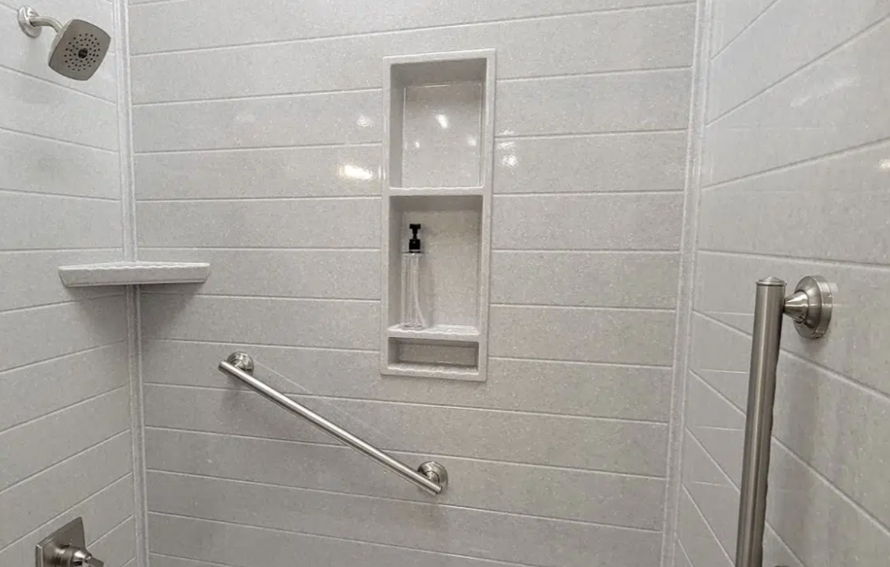 Shower area with white tiled walls, a showerhead in the upper left corner, a built-in shelf with a bottle of soap, and metal grab bars for support.