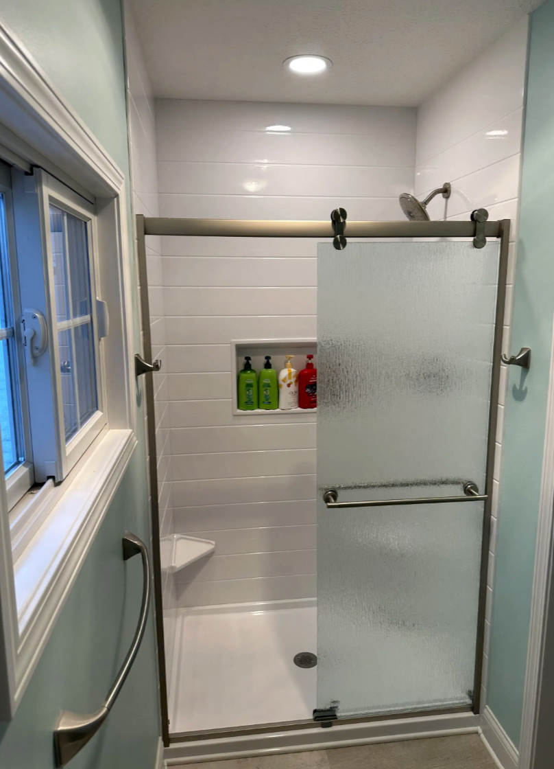 Small bathroom shower with frosted glass sliding door, white tiled walls, built-in shower caddy with shampoo and soap bottles, window with white frame, and light blue walls.