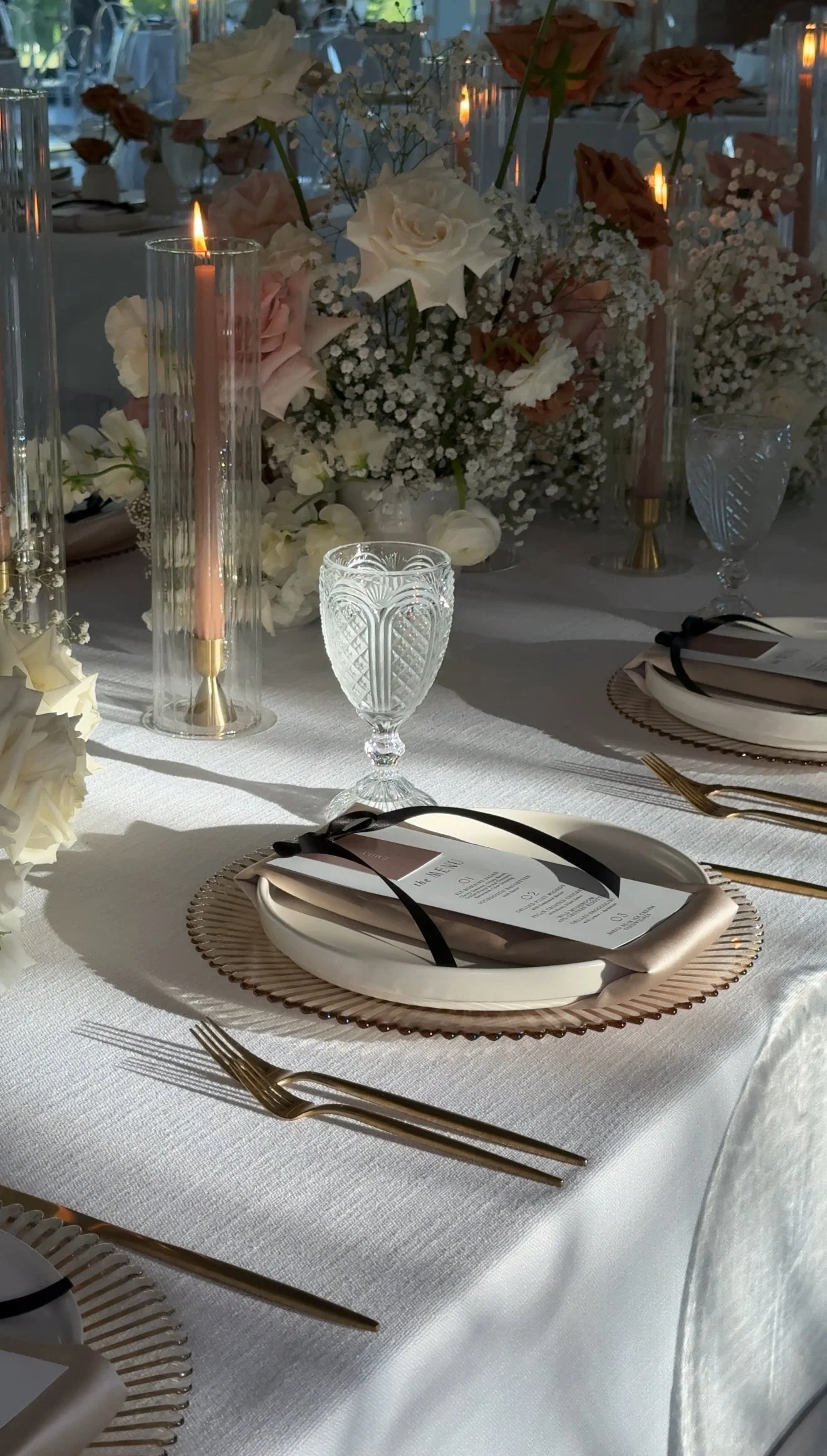 Elegant table setting with gold flatware, white plates on a pink scalloped charger, crystal glasses, pink candles, white napkin with black ribbon, and floral arrangements with white, blush, and brown flowers.