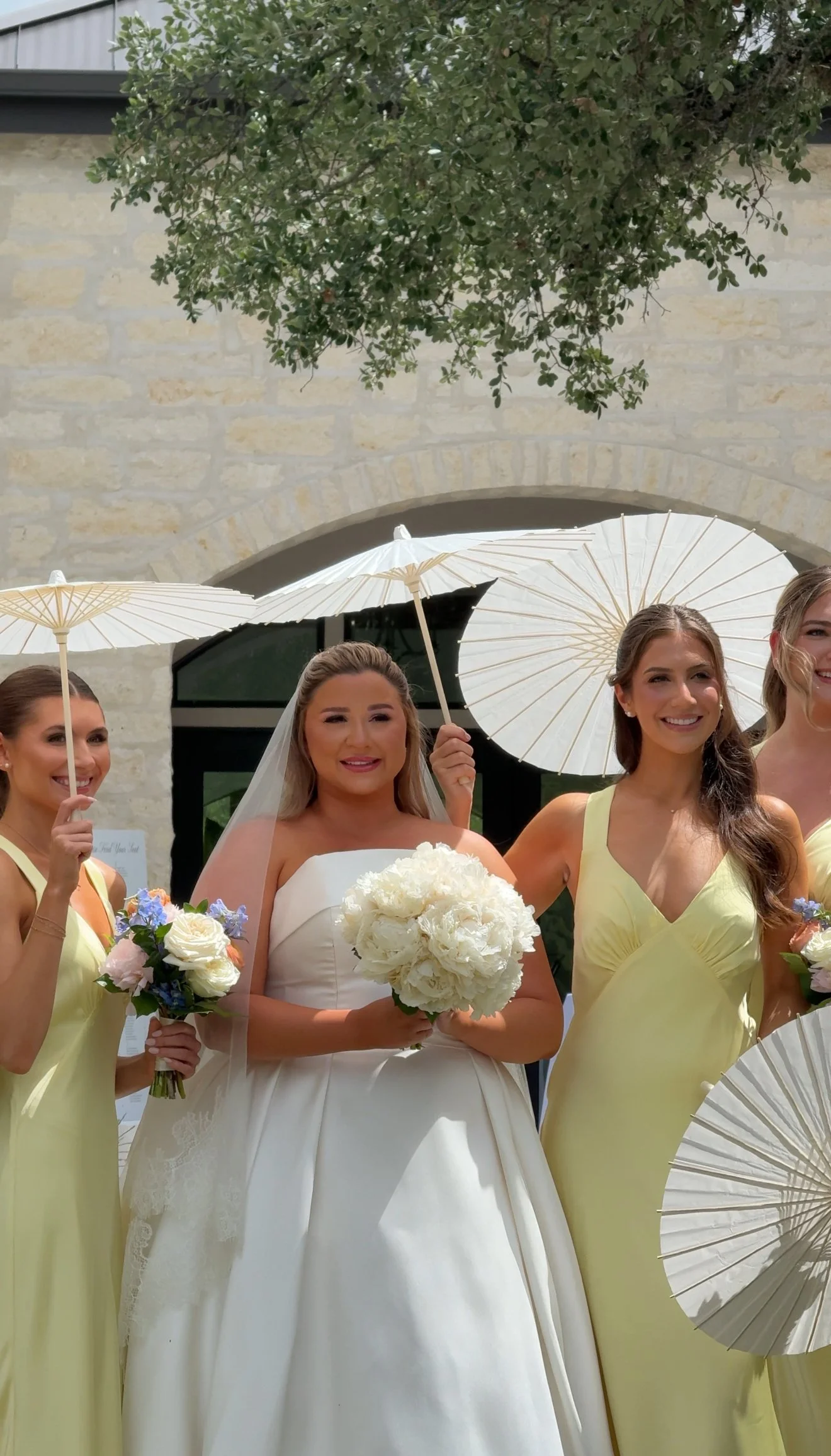 Wedding bride in white gown holding a bouquet, surrounded by bridesmaids in yellow dresses holding small bouquets and parasols, outdoors with stone building and greenery