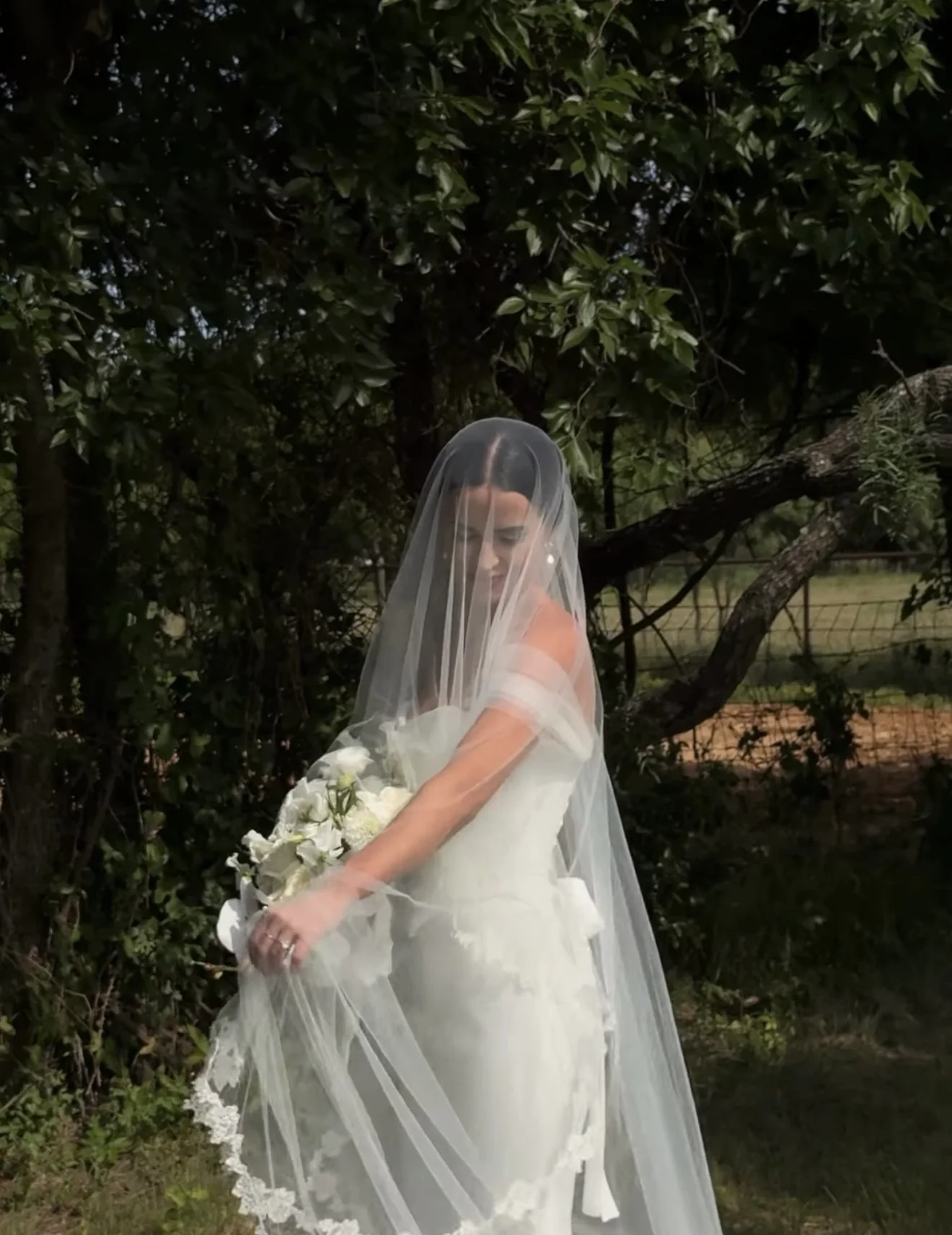 A bride in a wedding dress holding a bouquet of white flowers, standing outdoors behind a large tree, with her face partially obscured by a sheer veil.