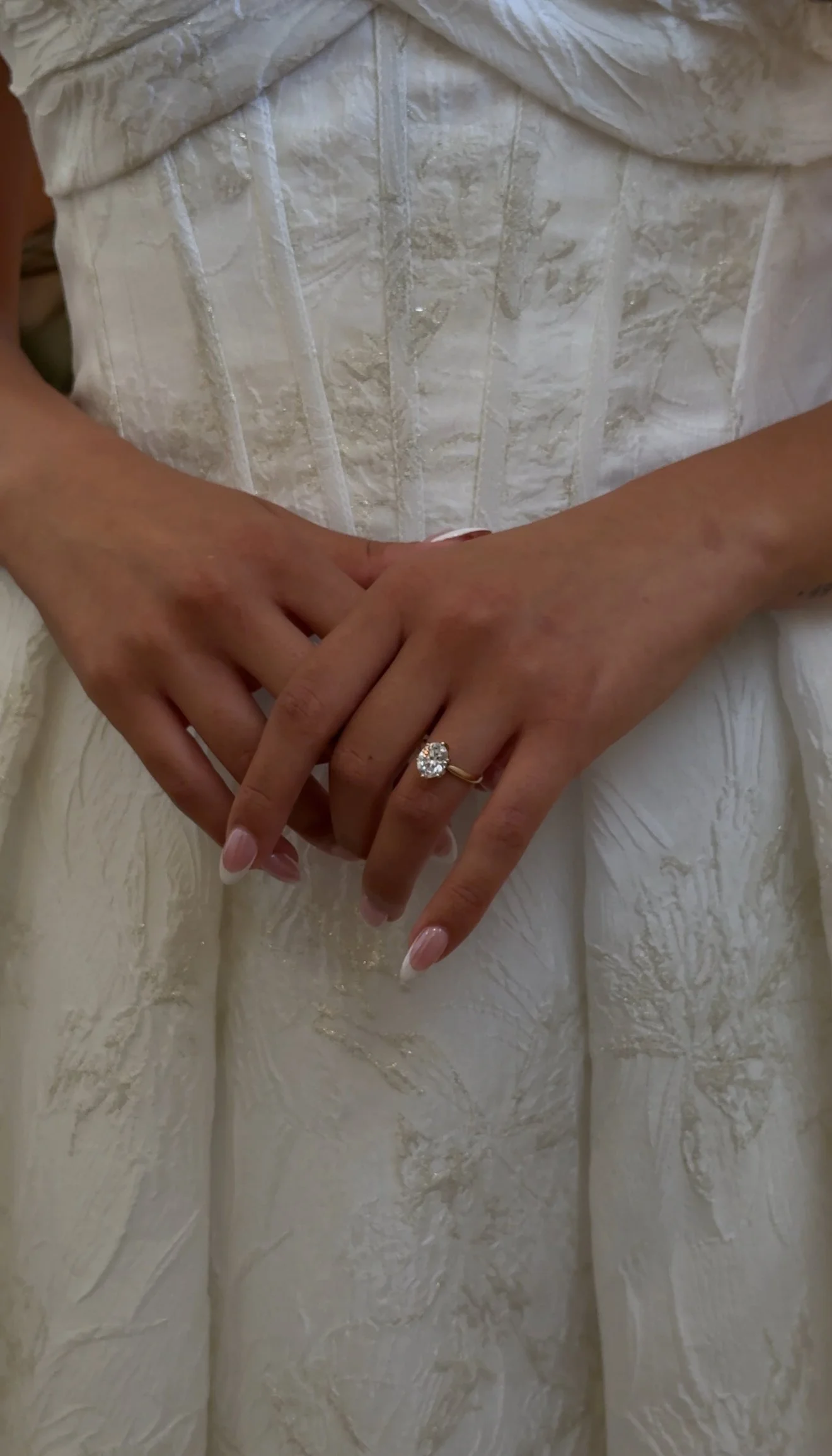 A bride in a white wedding dress showing her hands with an engagement ring with a large diamond, resting against her dress.